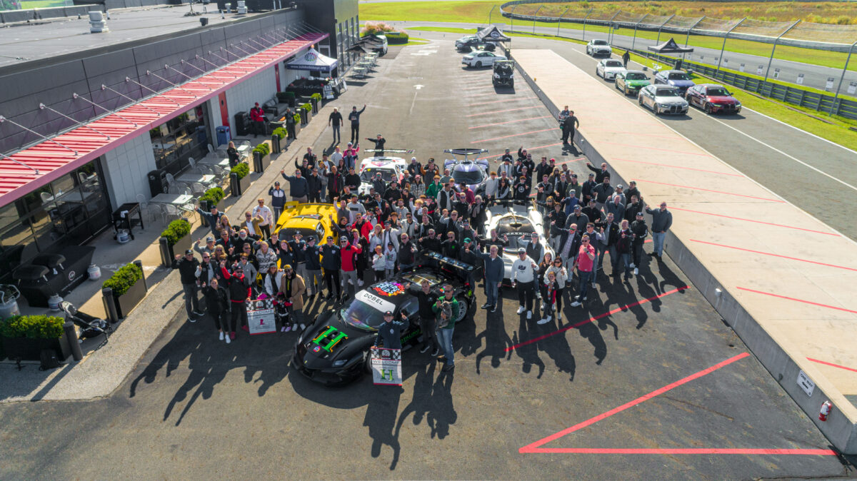 A large charity group posing around 5 race cars in pit lane